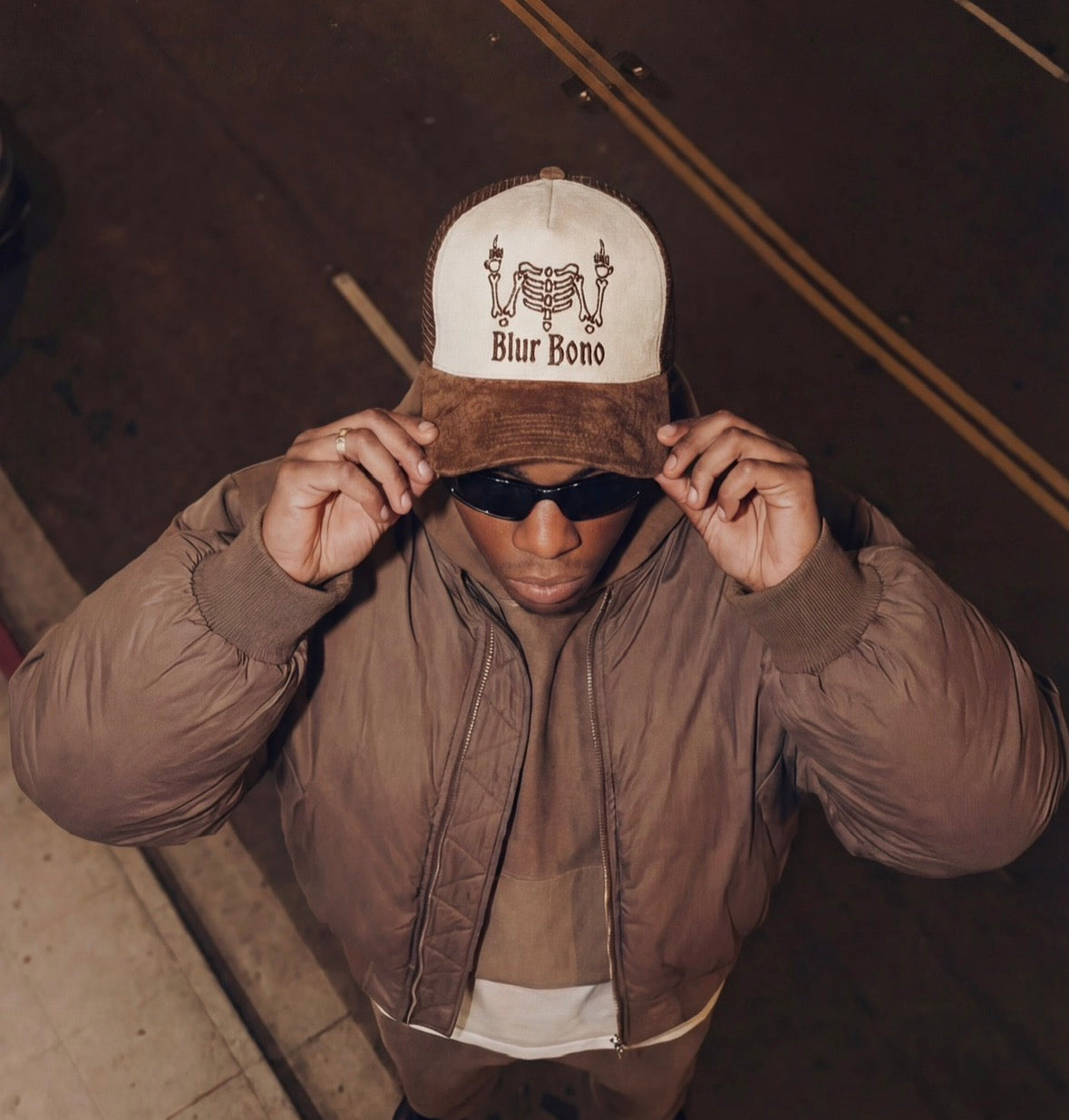 Bild tagen snett ovanifrån på man klädd i brun jacka och en brun och offwhite färgad keps med blur bono och ett skelett